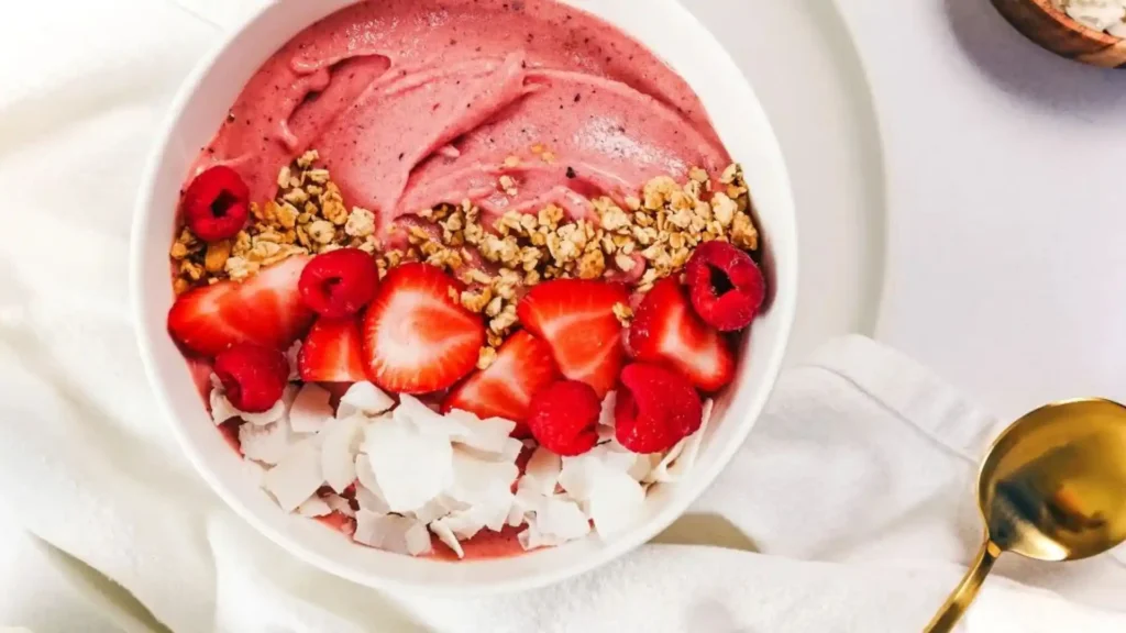 Pink smoothie bowl with raspberry blend, topped with fresh strawberries, raspberries, granola and coconut flakes, served with a gold spoon on white linen